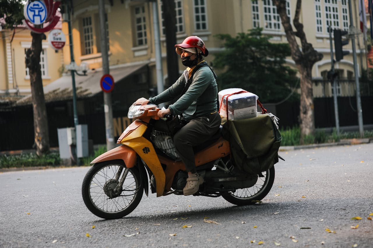 A motorcycle courier in a red helmet rides through an urban street, carrying a large package.